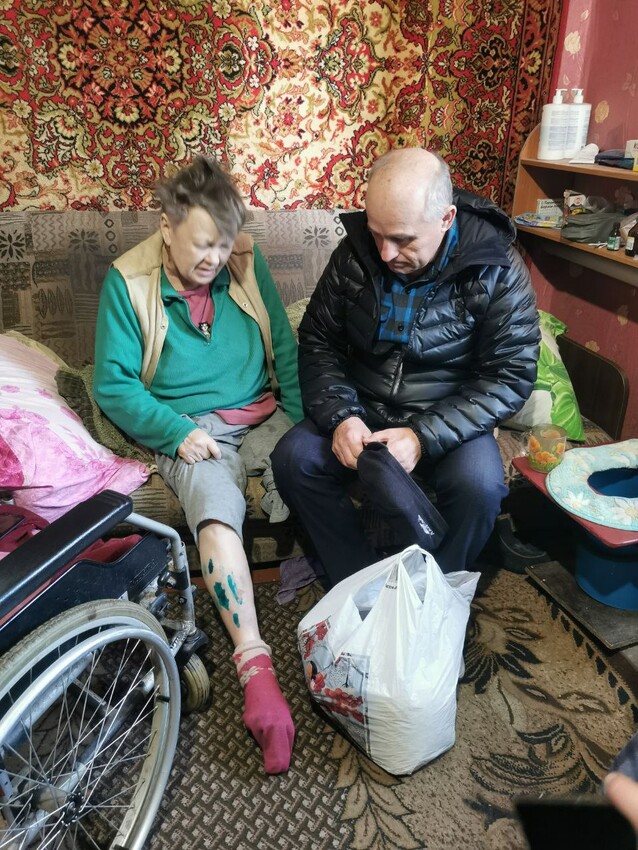 Two people sit on a couch. The person on the right is examining the person on the left's leg.