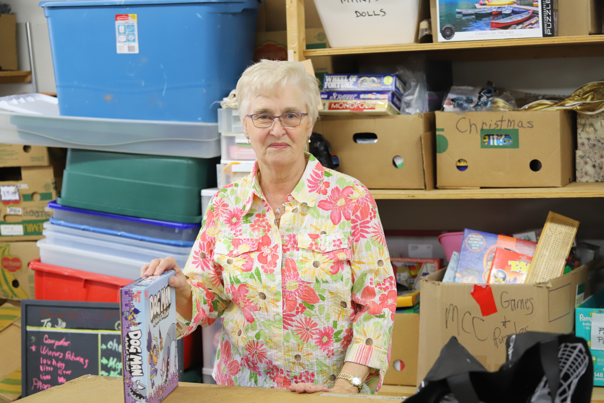 An older woman in a floral shirt stands in front of a wall of thrift shop donations