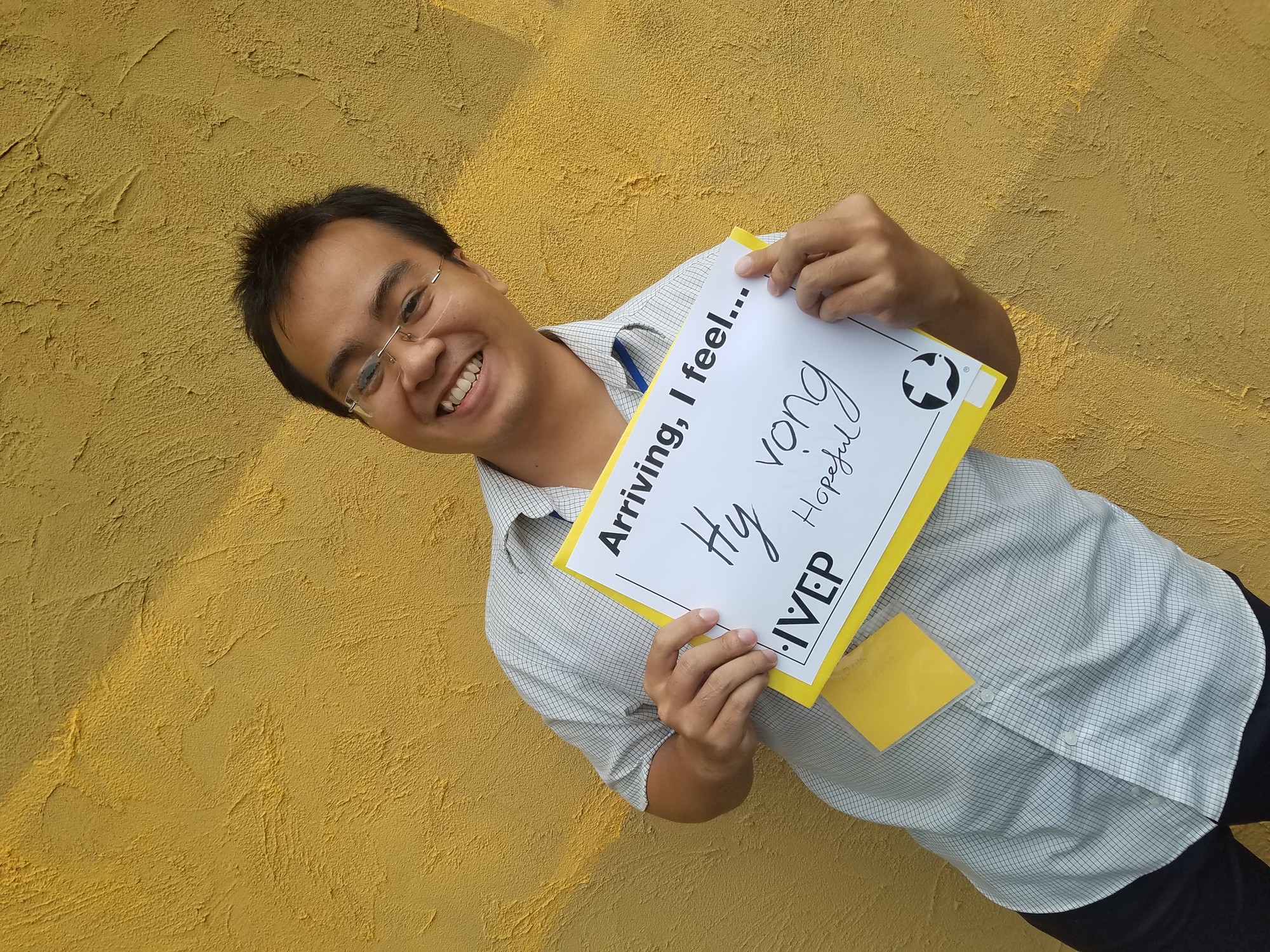 A young man from Vietnam stands in front of a yellow wall and holds a sign that says "Arriving I feel hopeful"