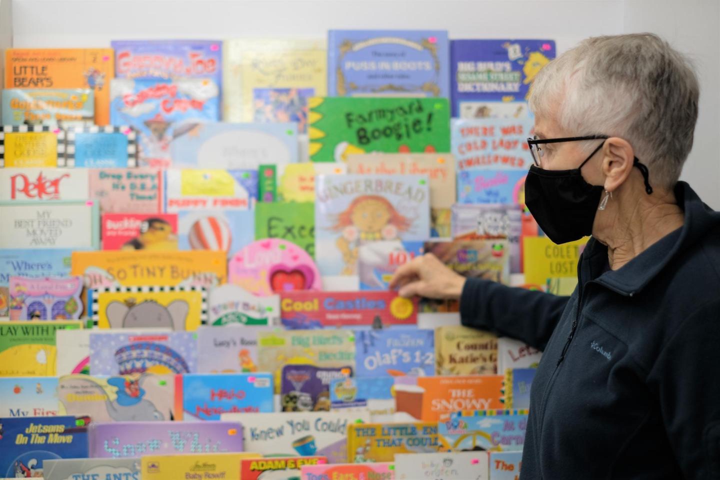 A woman with glasses and face mask touches a book in a book display at a thrift shop