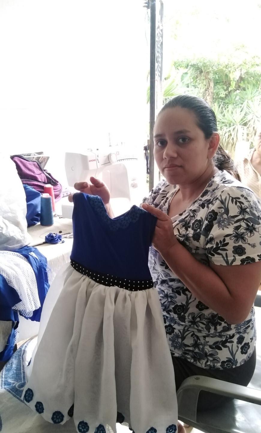 A woman sitting in front of a sewing machine holds up a child's dress.