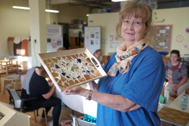 A woman in a blue shirt and a scarf around her neck holds up a mosaic tray