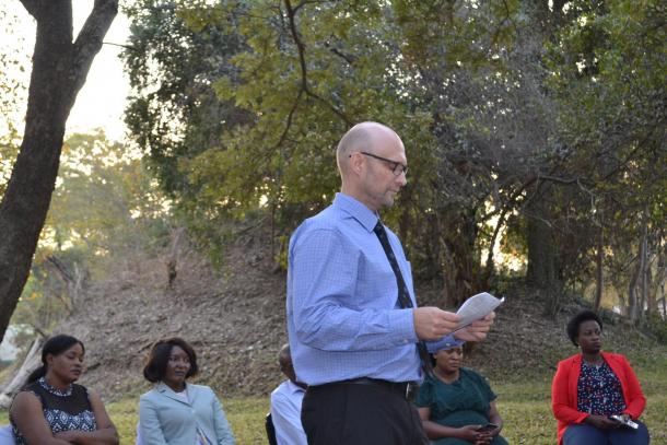 A bald man with glasses stands outside and reads something from a piece of paper.