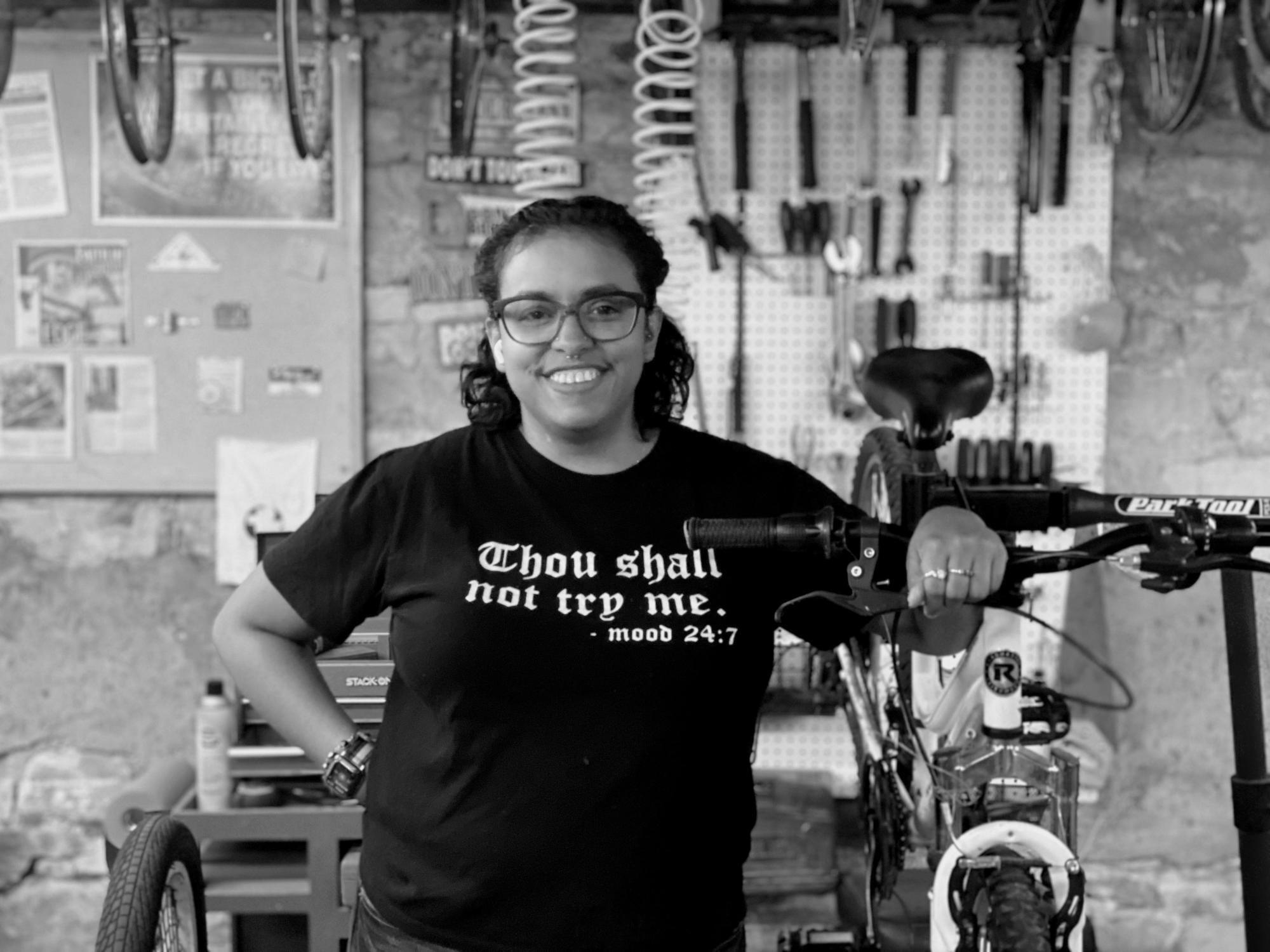 A woman stands next to a bike. Behind her is a wall of bike parts.