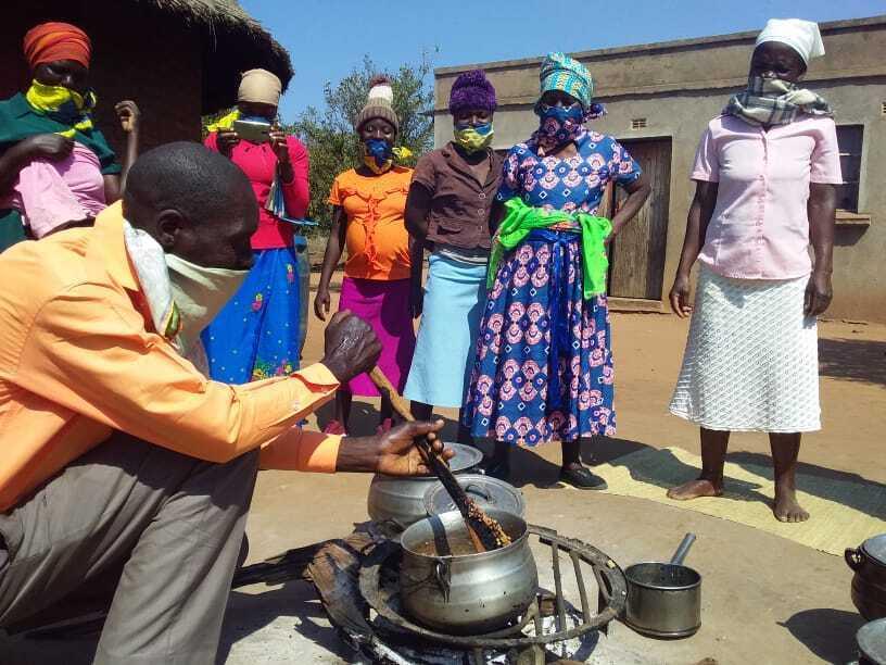 A man squatting in front of an outdoor fire. He is stirring something in a pot. There are six woman watching him.