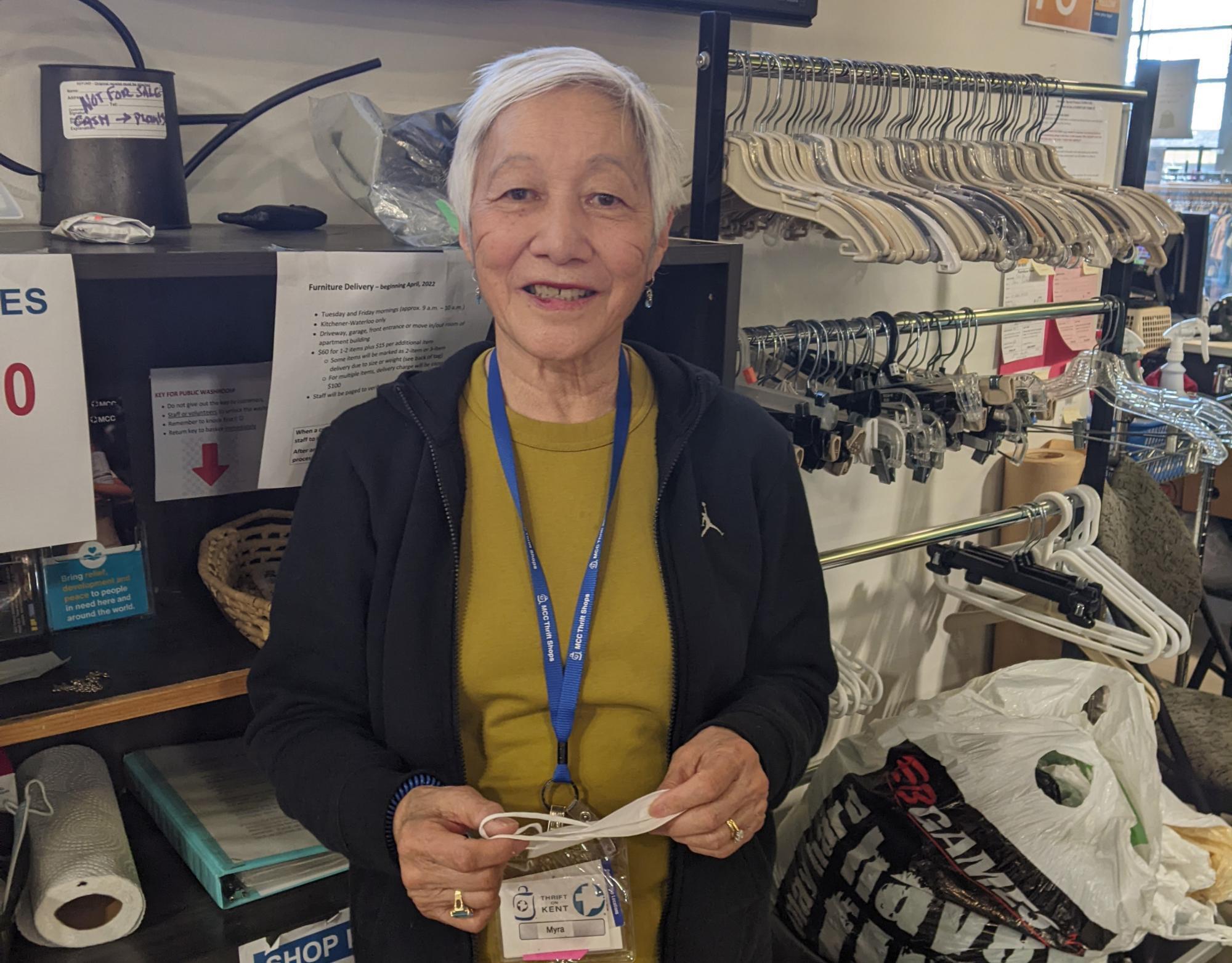 An old woman in a black jacket and green shirt smiles for the camera. She is standing in front of a rack of clothes hangers. 