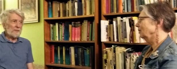 An older man and woman sit facing each other in front of a shelves of books