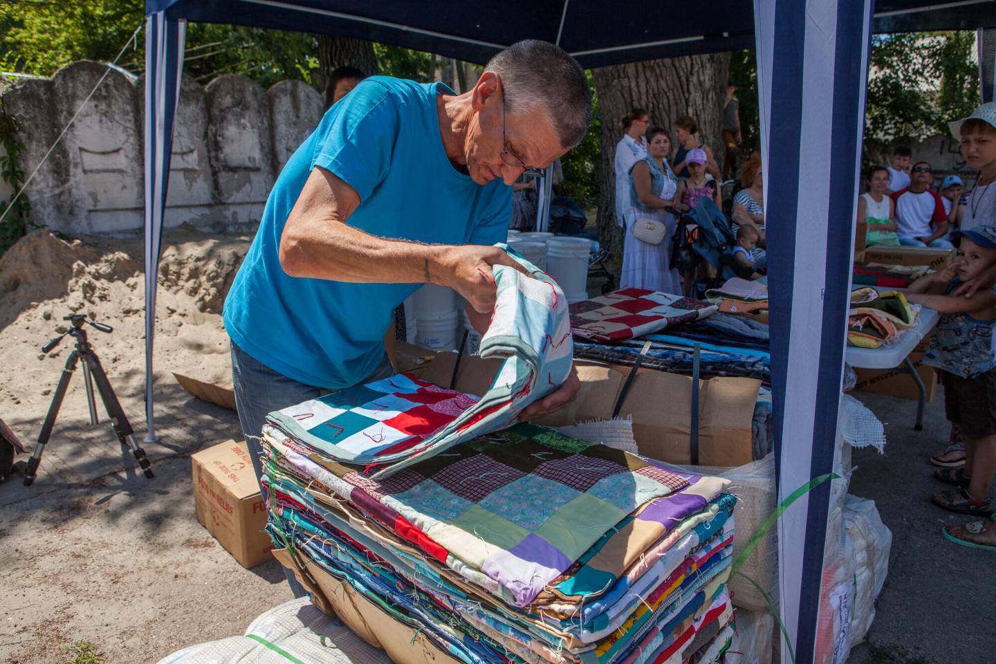 A man in a blue shirt and glasses lifts a colorful comforter off a pile of blankets