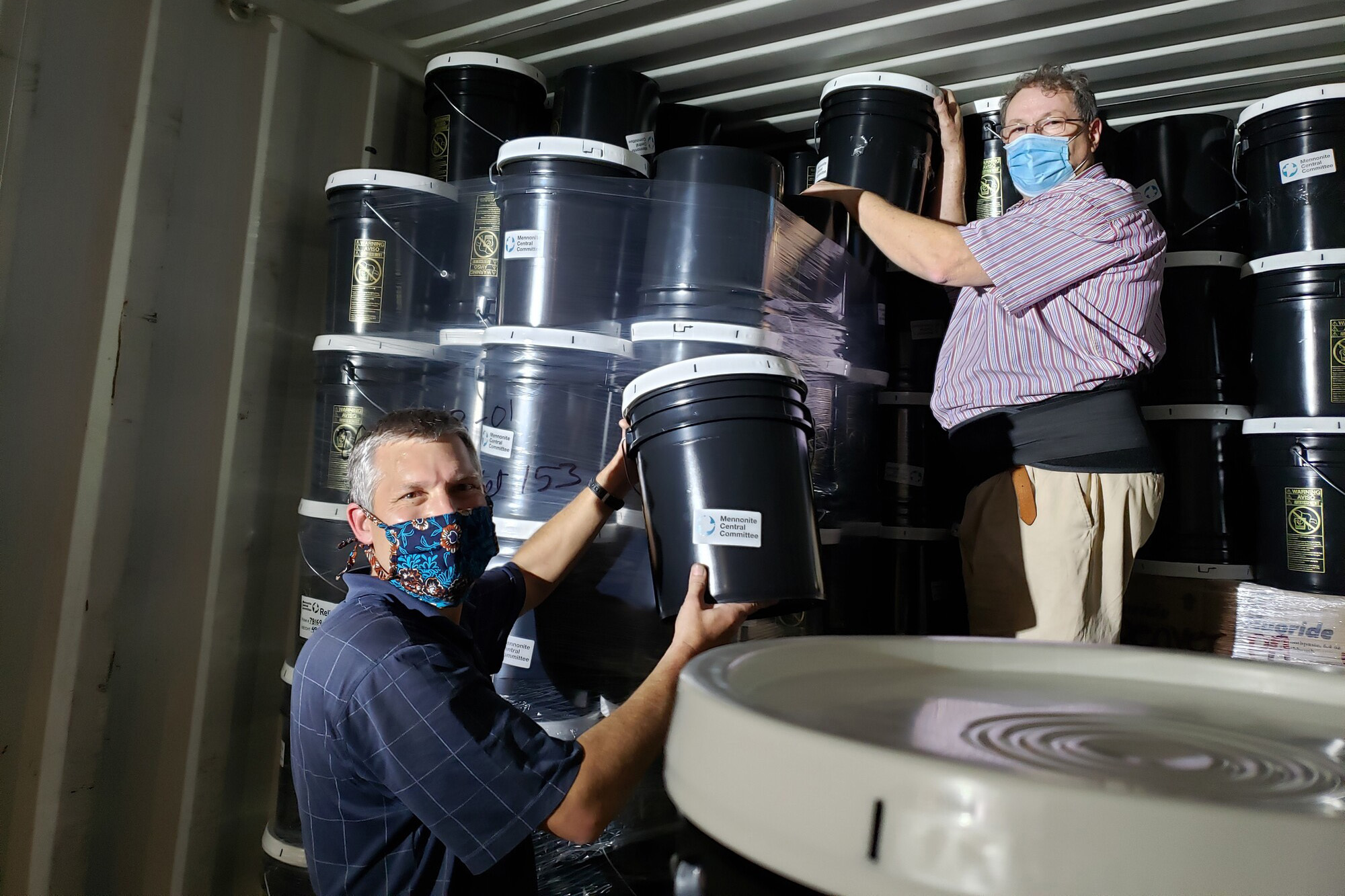 Two men load buckets into a shipping container. They both are looking at the camera.