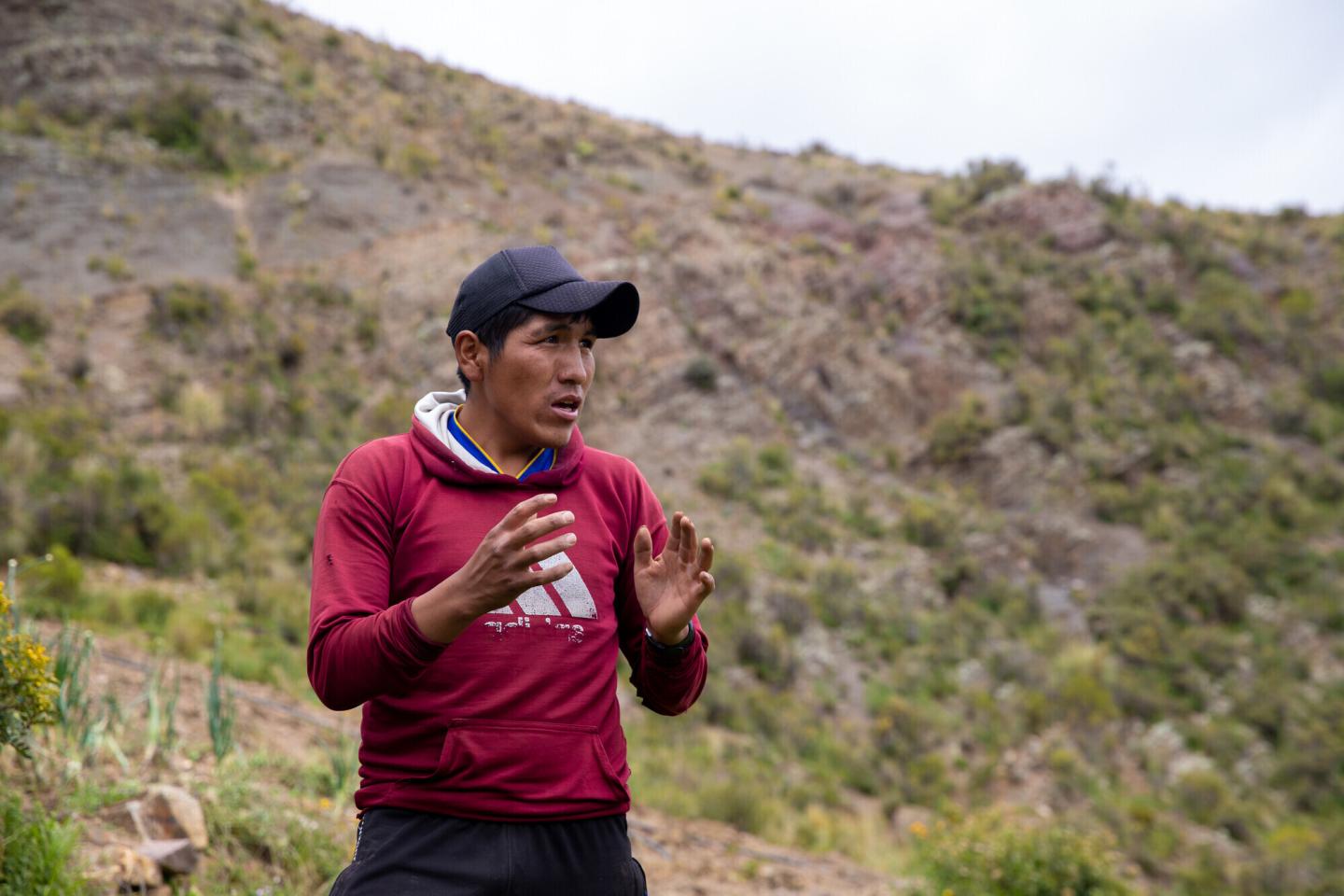 A Bolivian man in a red sweatshirt and black cap speaks and gestures with his hands