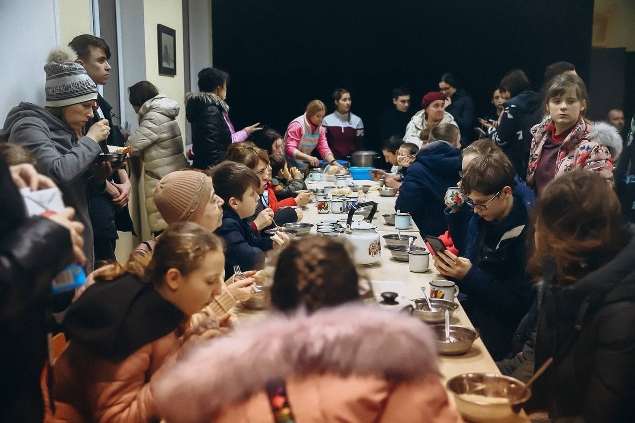 A large group of people in coats and winter hats gather around a table and eat