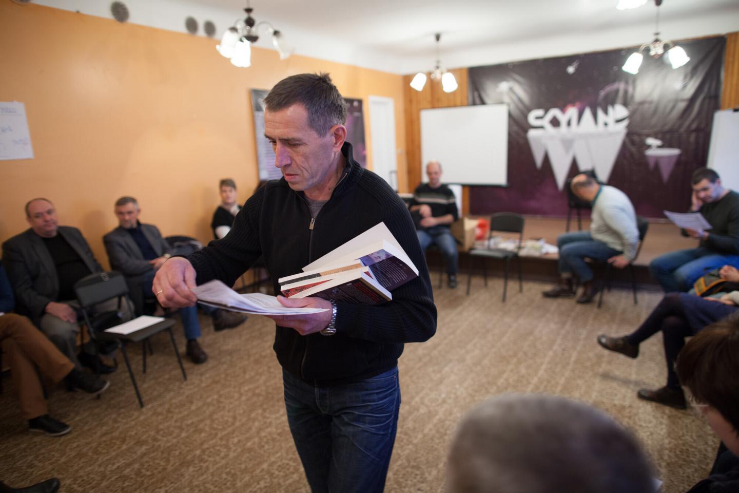 A man holding a pile of books passes out pieces of paper to a group sitting around him in a circle