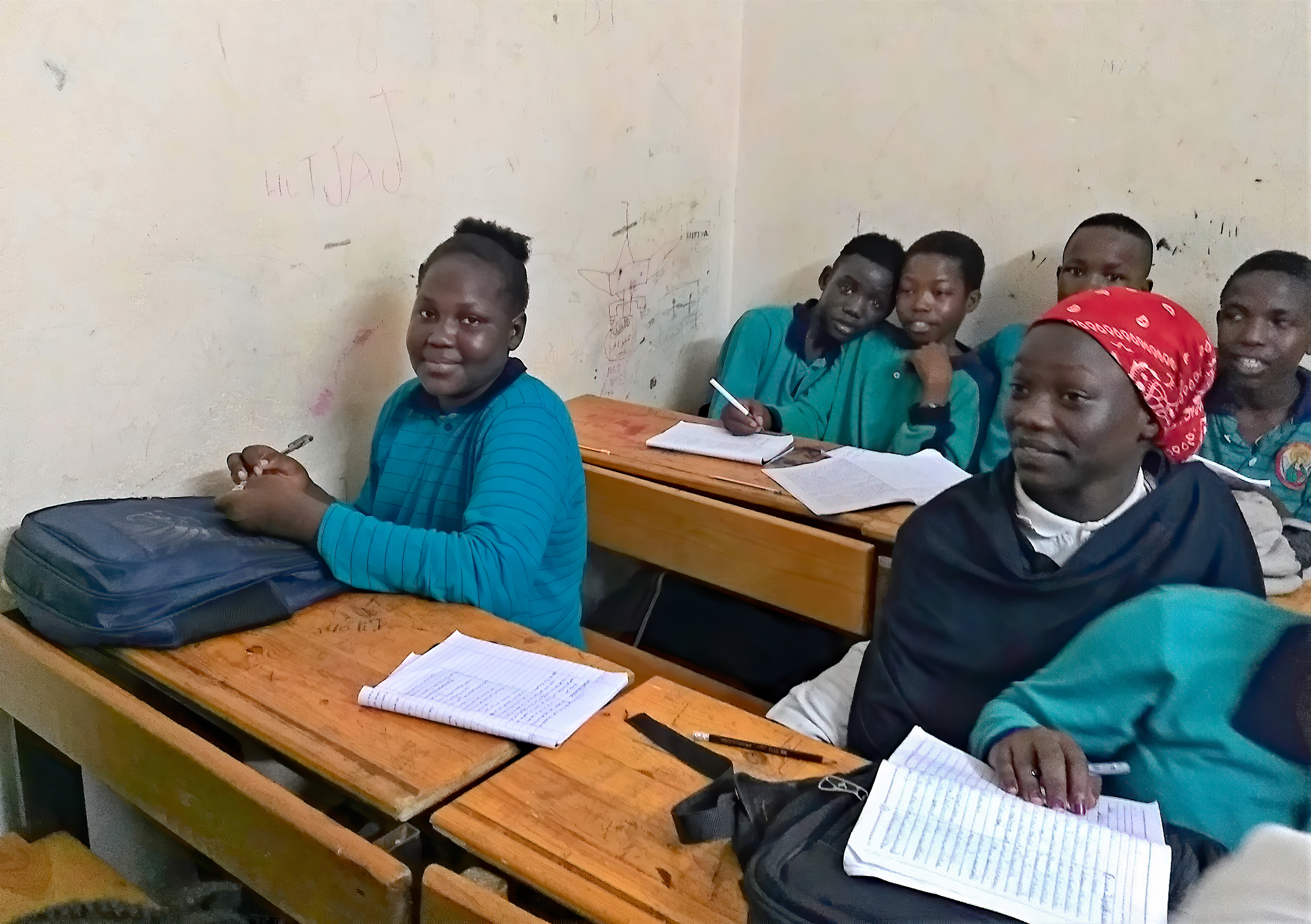 A group of Sudanese students sit at their desks in a small classroom.