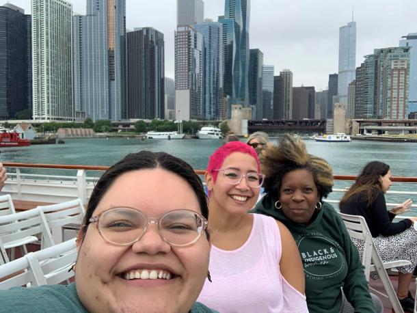 Three women on a boat deck that is floating in a river surrounded by skyscrapers.