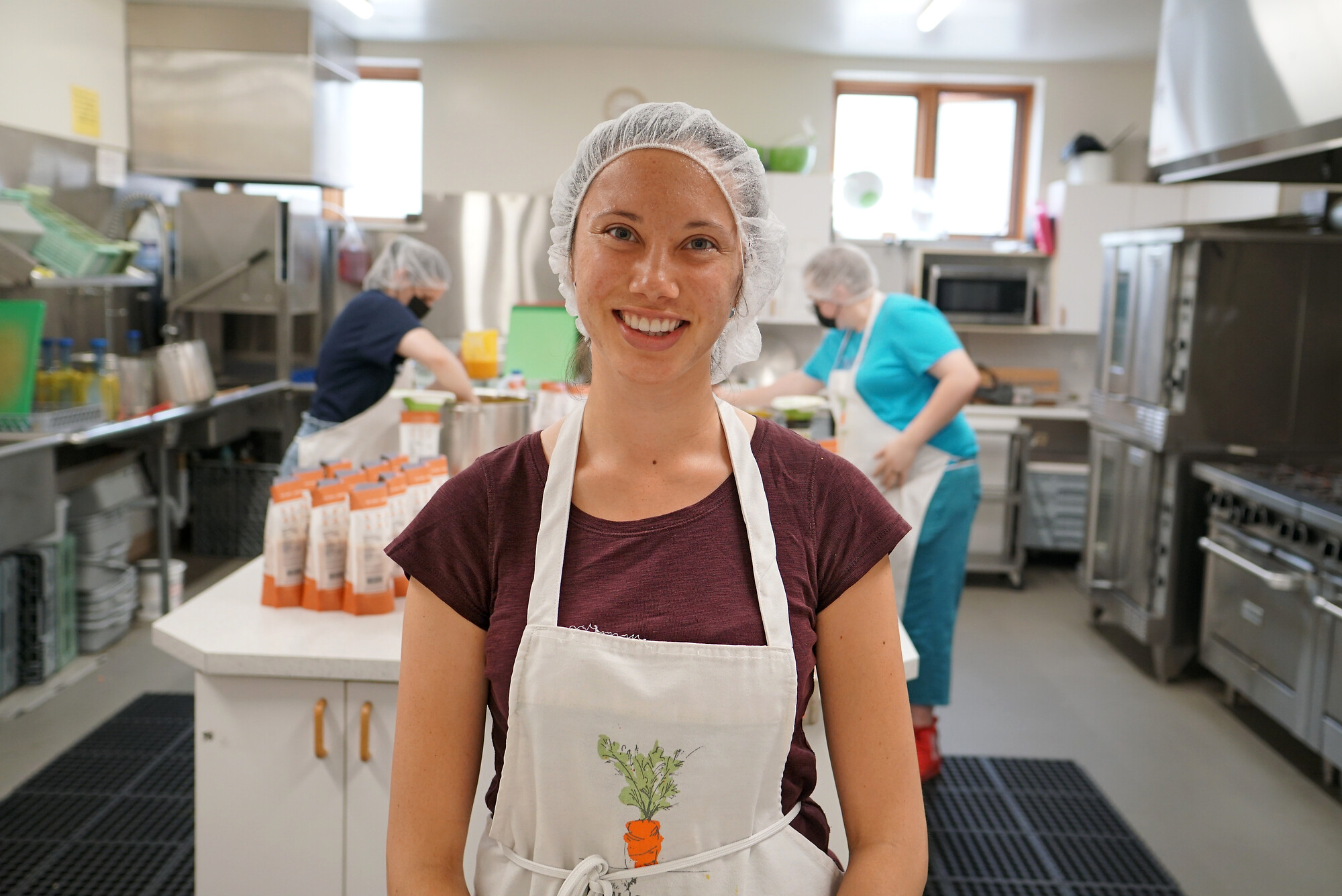 A woman in a hair net and apron stands in a kitchen. Behind two people in hair nets, face masks and aprons work.