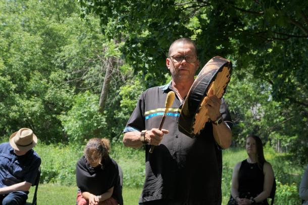 A man stands outside and pounds on a hand held drum. There are people sitting behind him, listening.