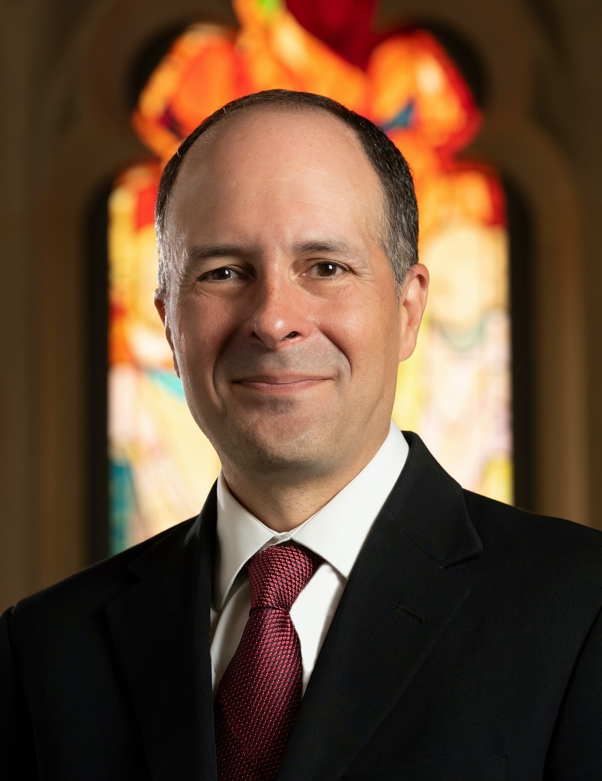 A headshot of a man in a suit and tie standing in front of a stain glass window