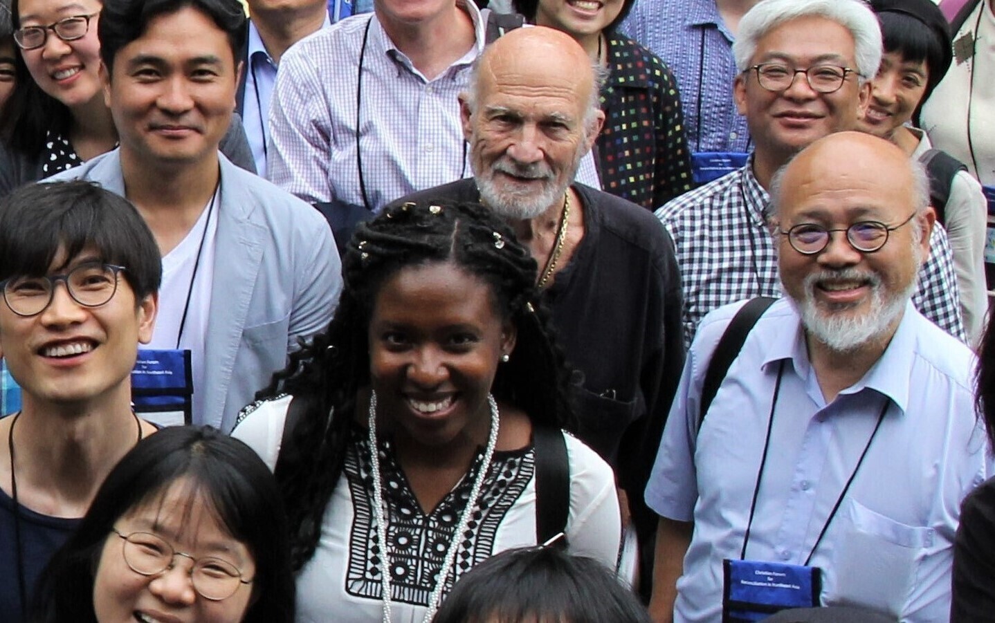 A view from above of a large group of adults made up of many different ethnicities. A bald man with a white beard wearing a black top stands in the middle.