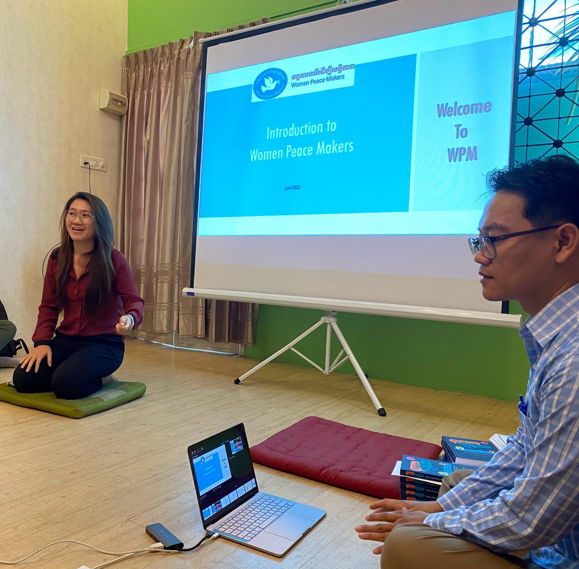 Two people sit on the floor in front of a projector screen. Words on the screen reads, "Introduction to women peace makers."