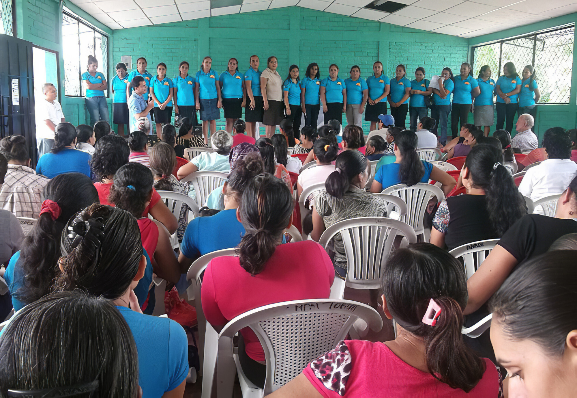 A large group of woman stand in a long line on a stage. There is a man with a microphone. There is also a large audience sitting in front of them.