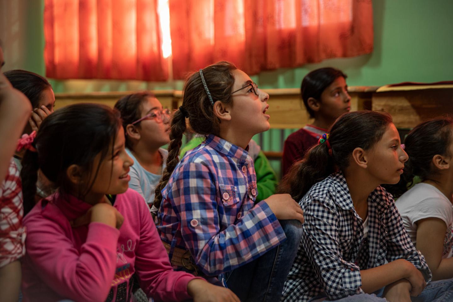 A group of school children sit and listen to someone out of frame on the right.