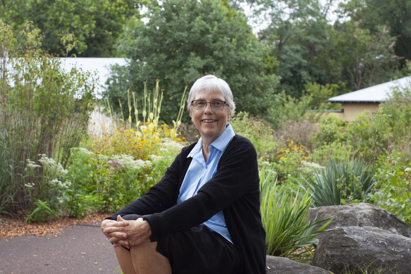 An older woman sitting on a large rock, surrounded by native plants and flowers