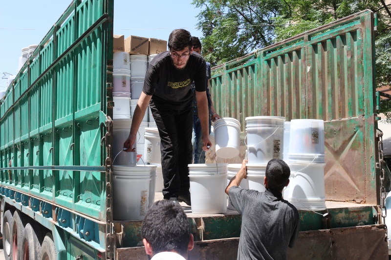 A person in the back of a trailer truck unloads five-gallon buckets to men standing on the ground.