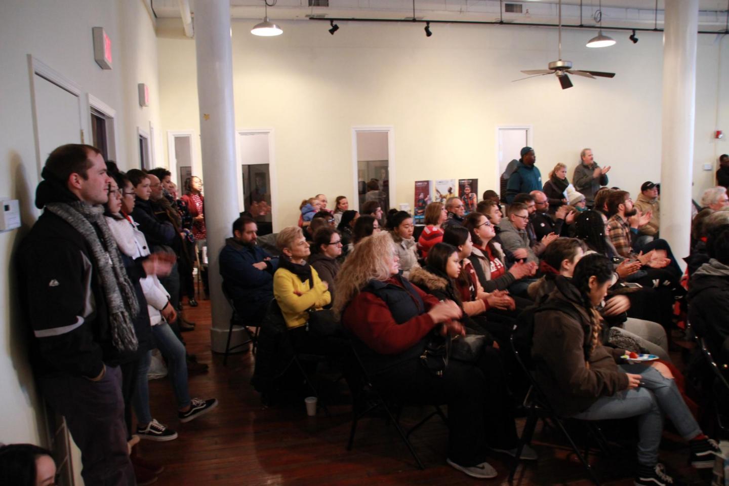 A large room of people sitting and standing listen to a speak that is off on the right.