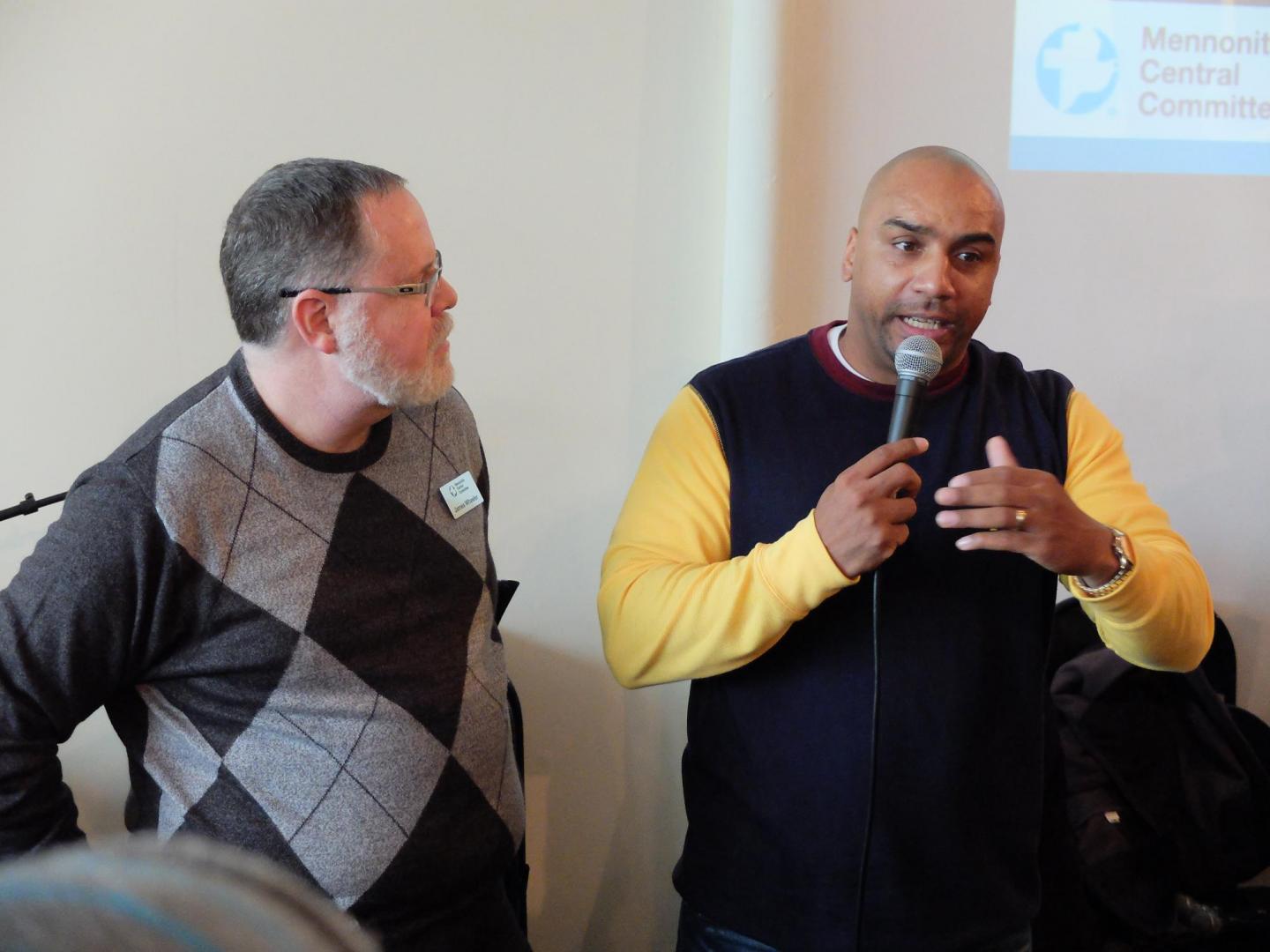 Two men stand next to each other. The man on the left is looking at the man on the right who is speaking into a microphone.