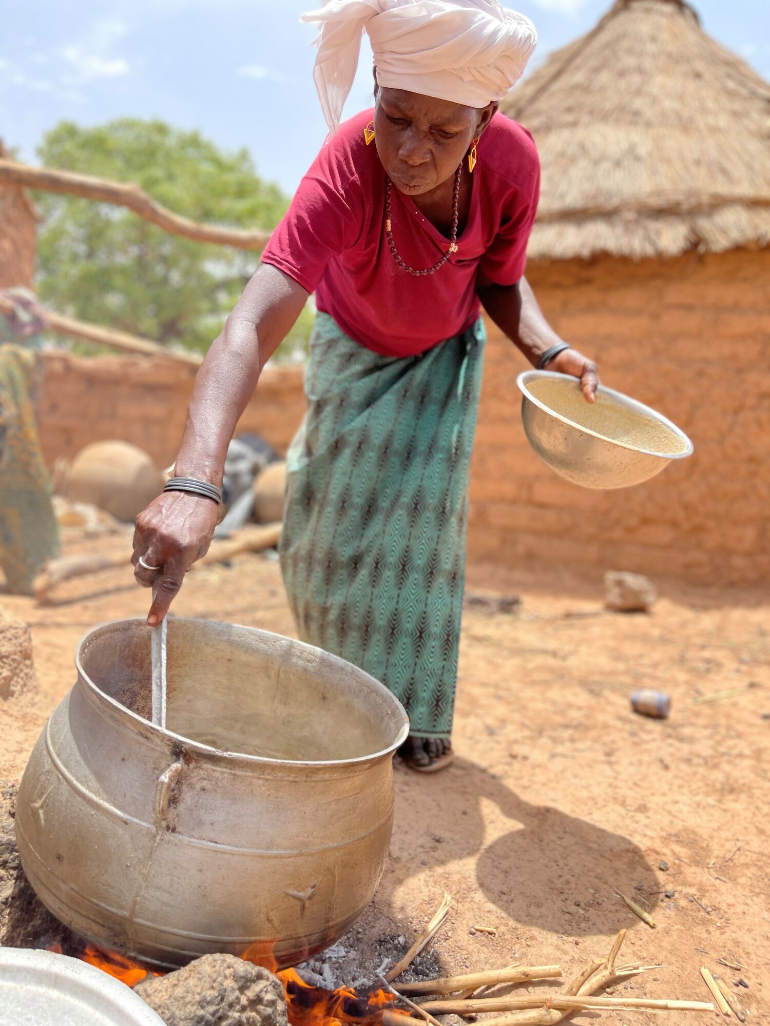 A woman leans over a large po cooking on an outside fire. She is holding a large spoon in one hand and bowl in the other