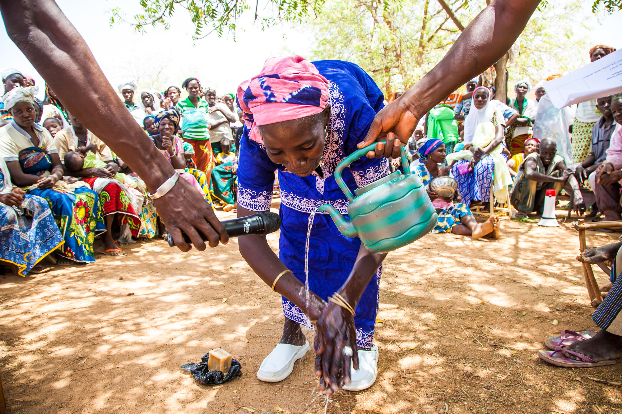 A woman demonstrates how to wash her hands. Someone is pouring water on her hands from a green tea pot while another person holds a microphone