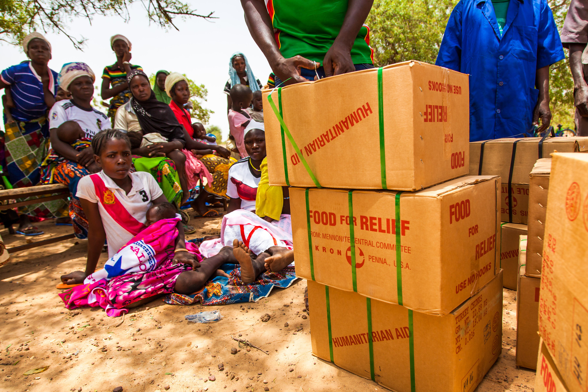 A group of people wait for as someone opens a stack of boxes that read, "food for relief"