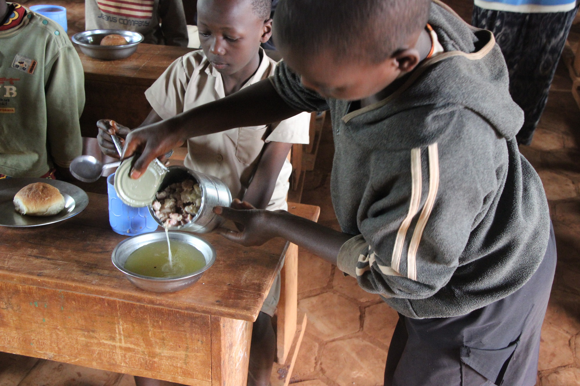 A Burundi student pours broth out of a can of meat into a bowl on a desk where other children are eating