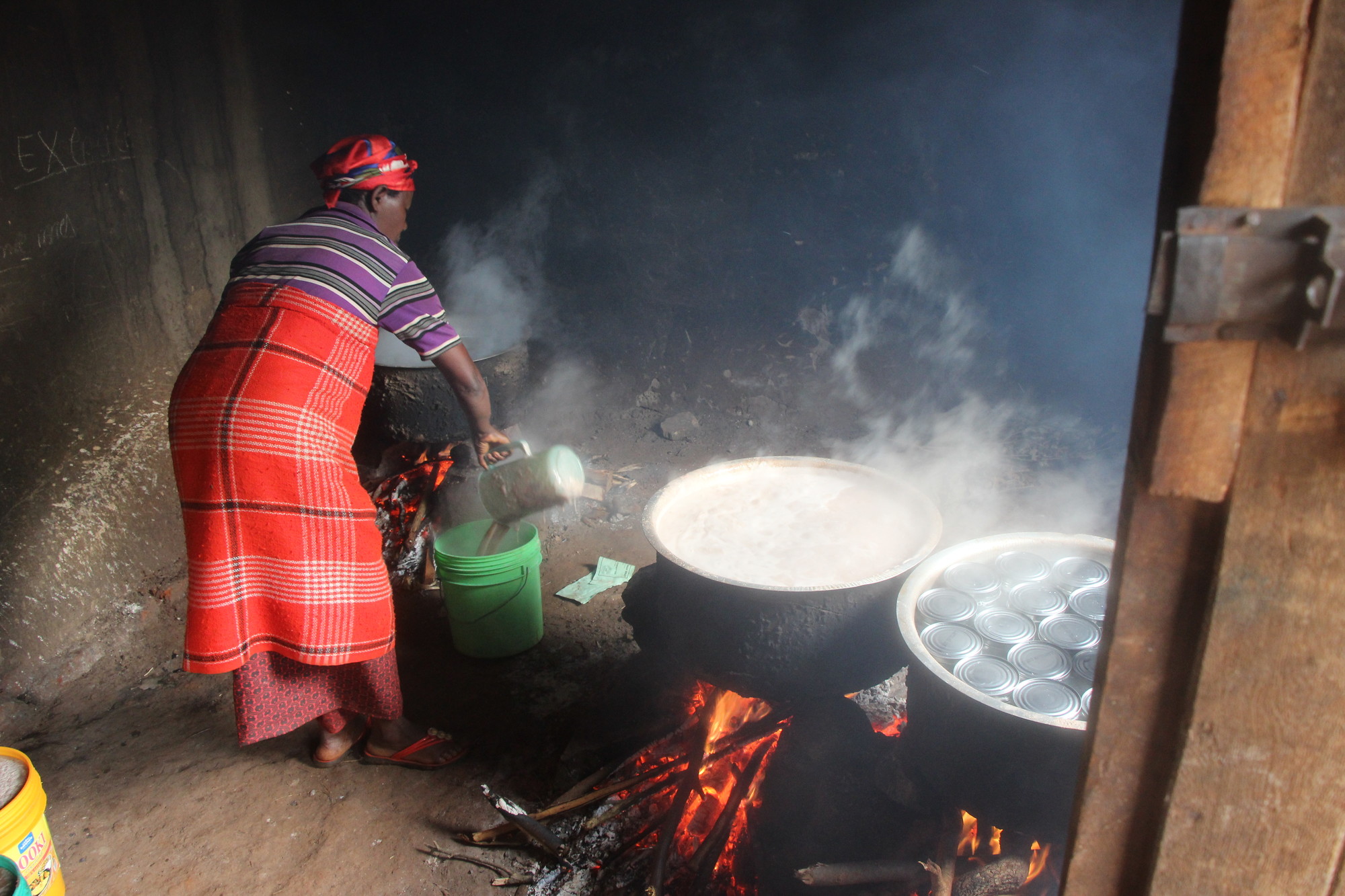 A Burundi woman poors liquid into a green bucket. Beside her are three large pots cooking over open fires