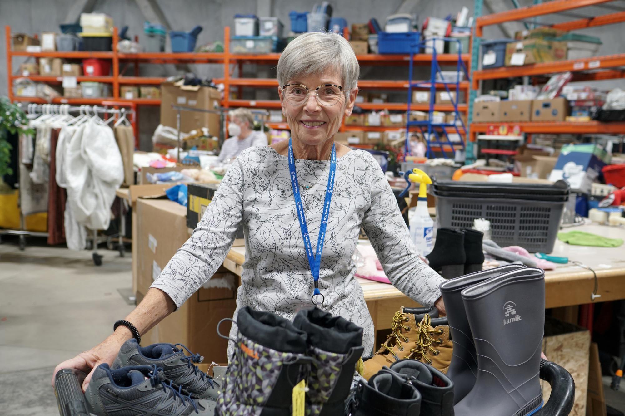 Woman with short silver hair and glasses stands behind a tray of boots and shoes. In the background is a warehouse with clothing and other items.