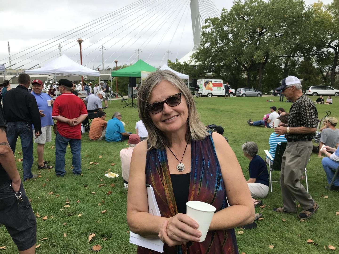 A  woman in sunglasses, with papers tucked under her arm, holds a paper cup. 