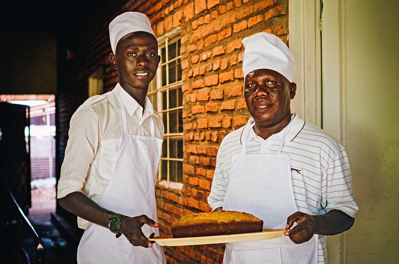 Two Rwandan chefs in white aprons and caps hold a try of banana bread