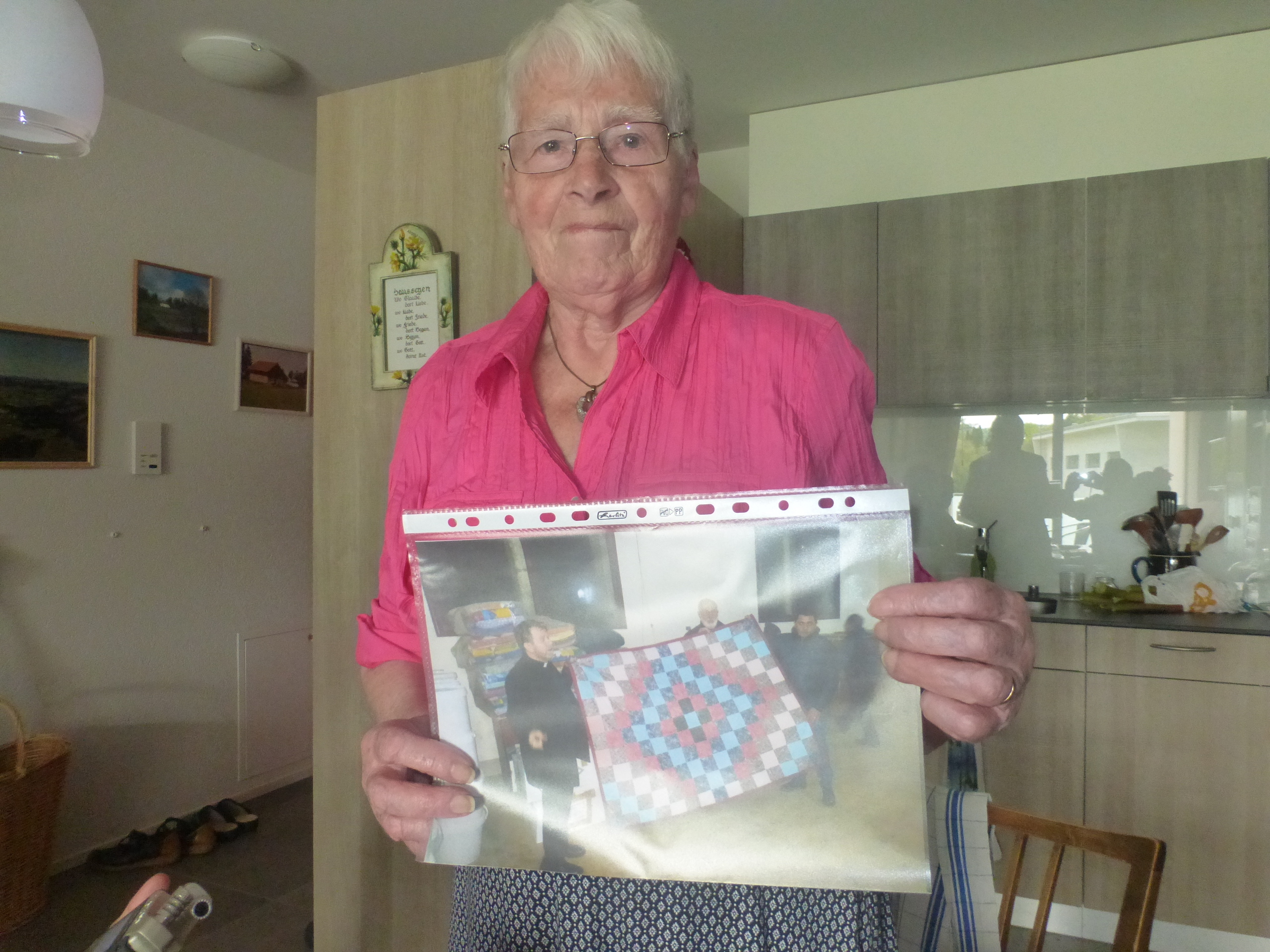 An older woman in a pink shirt holds a photo of people displaying a comforter