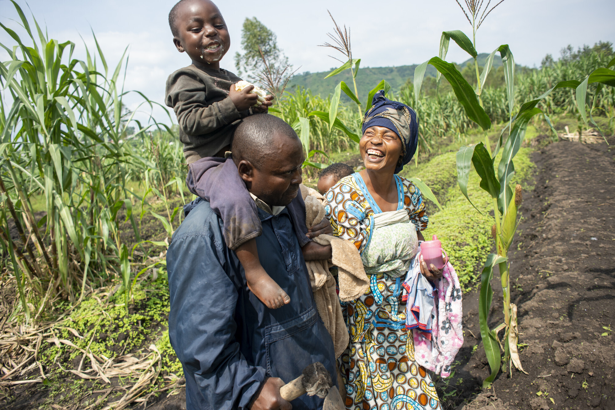 A family of four in the DR Congo walk together in a field. A child is on the father's shoulders laughing and eating