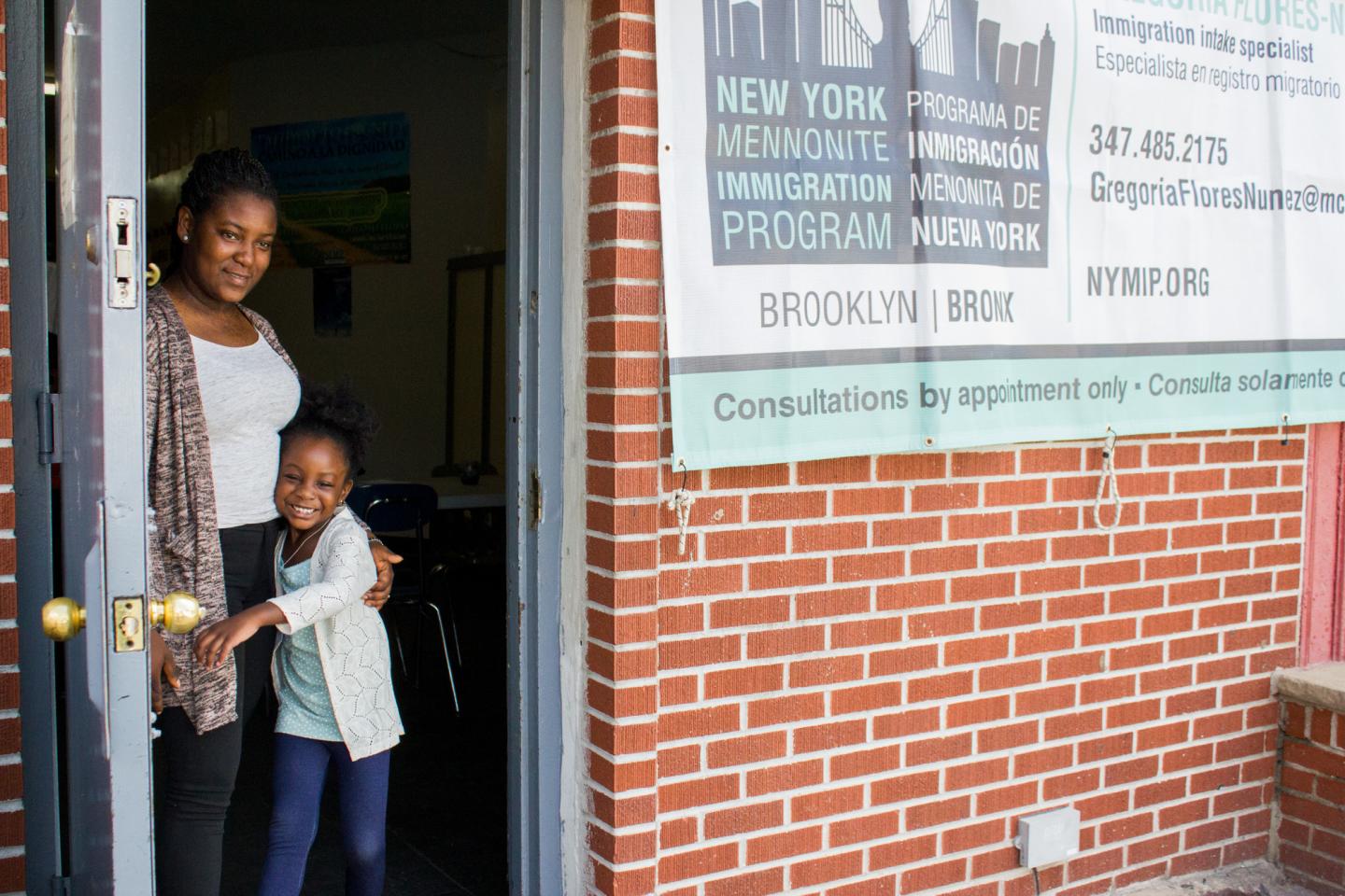 A mother and daughter stand in a doorway of a brickbuilding. The daughter is hugging the mother