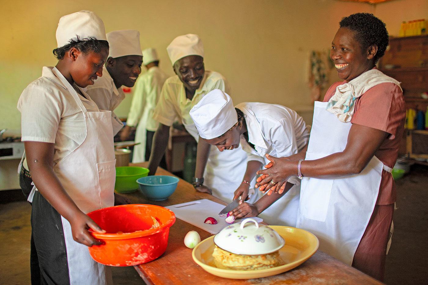 A group of culinary students in white aprons and chef hats watch a student chop an onion