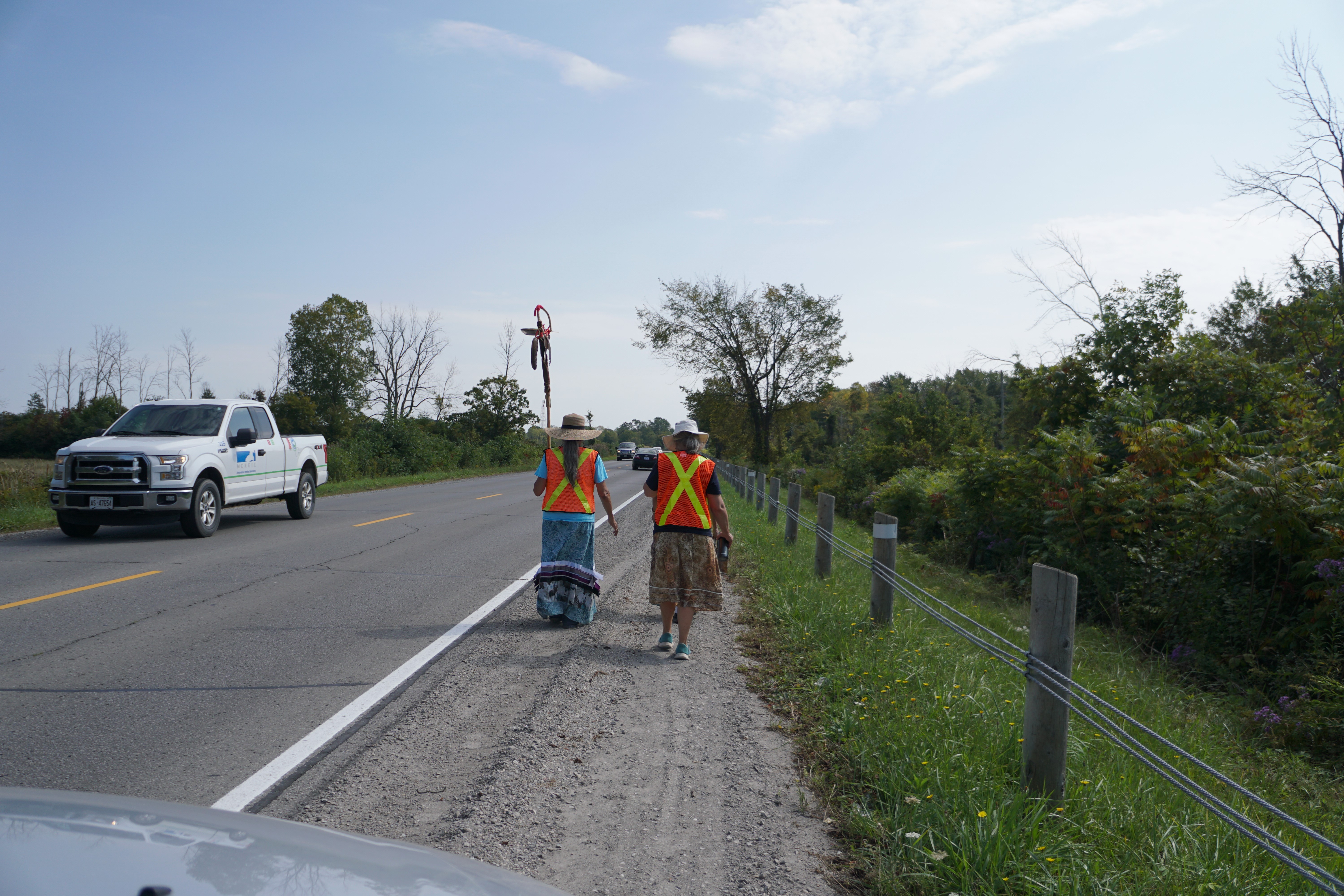 two women walk on the shoulder of the road as a truck passes by