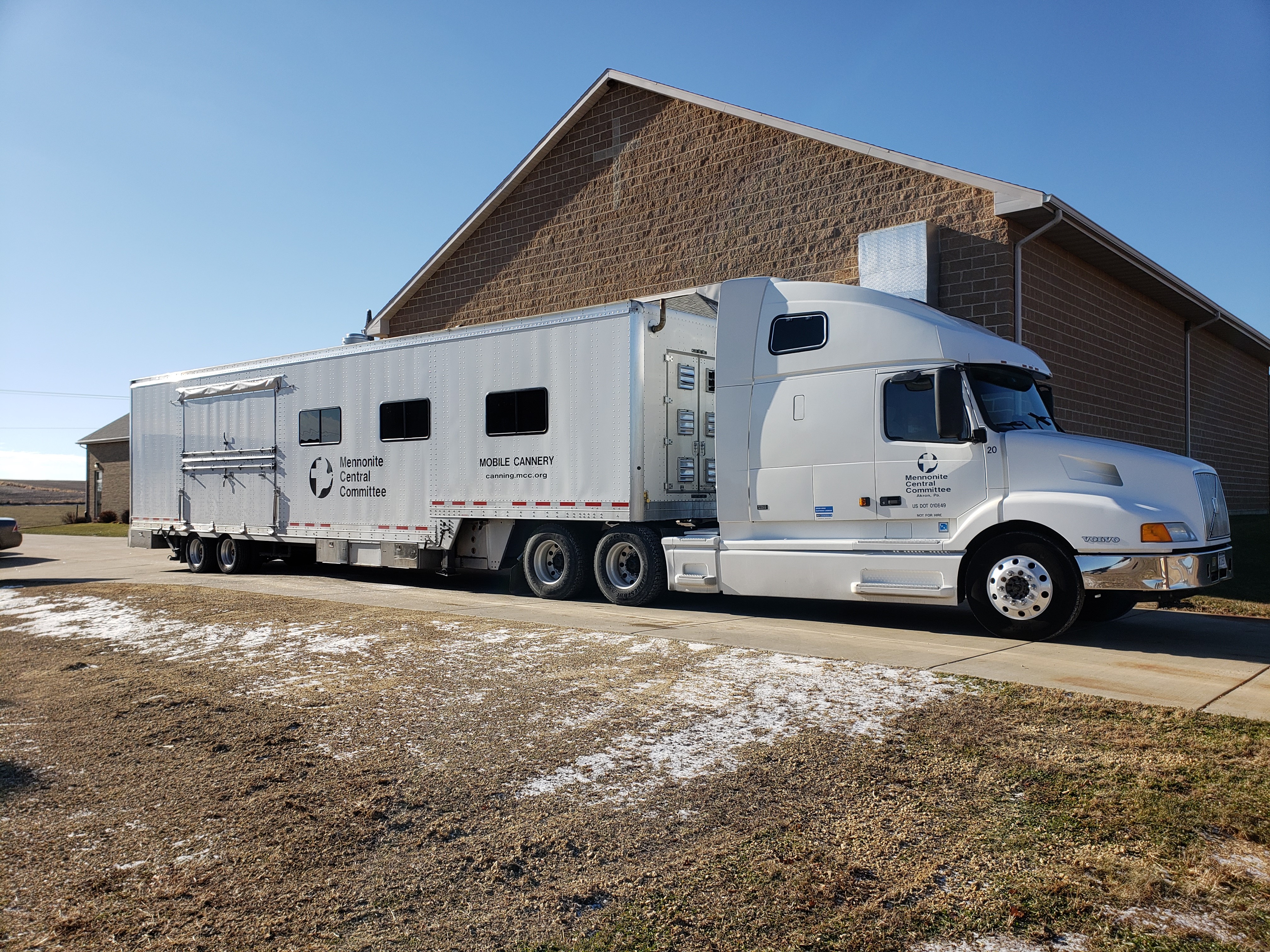 A long white tractor trailer rig with MCC logo in front of a church building