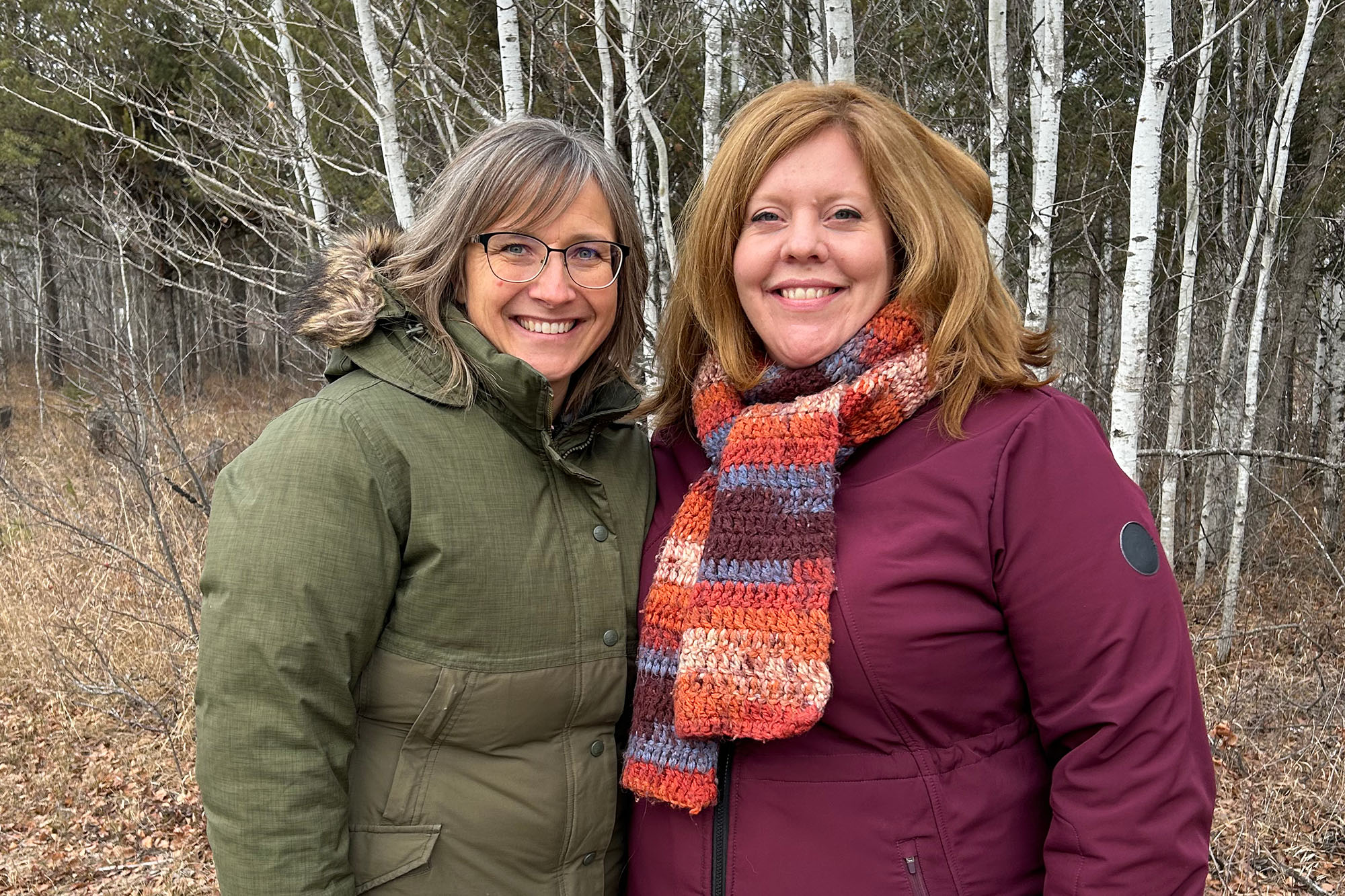 Two women standing in a forest and smiling at the camera.