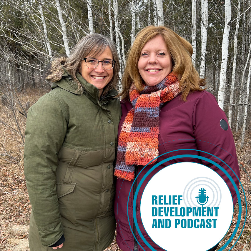 Two women standing in a forest and smiling at the camera.