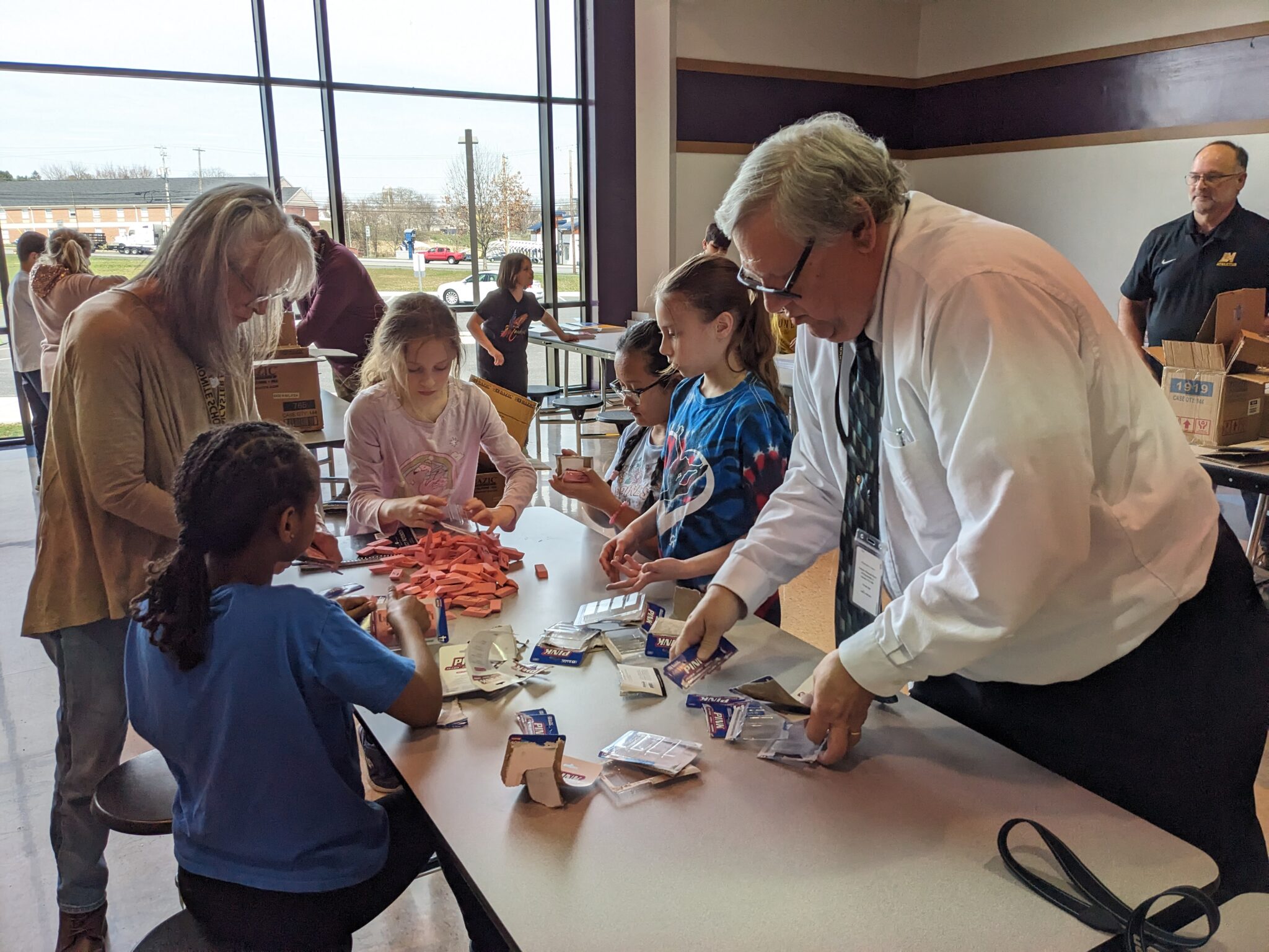 Three students and two teachers gather around a table with supplies for school kits.