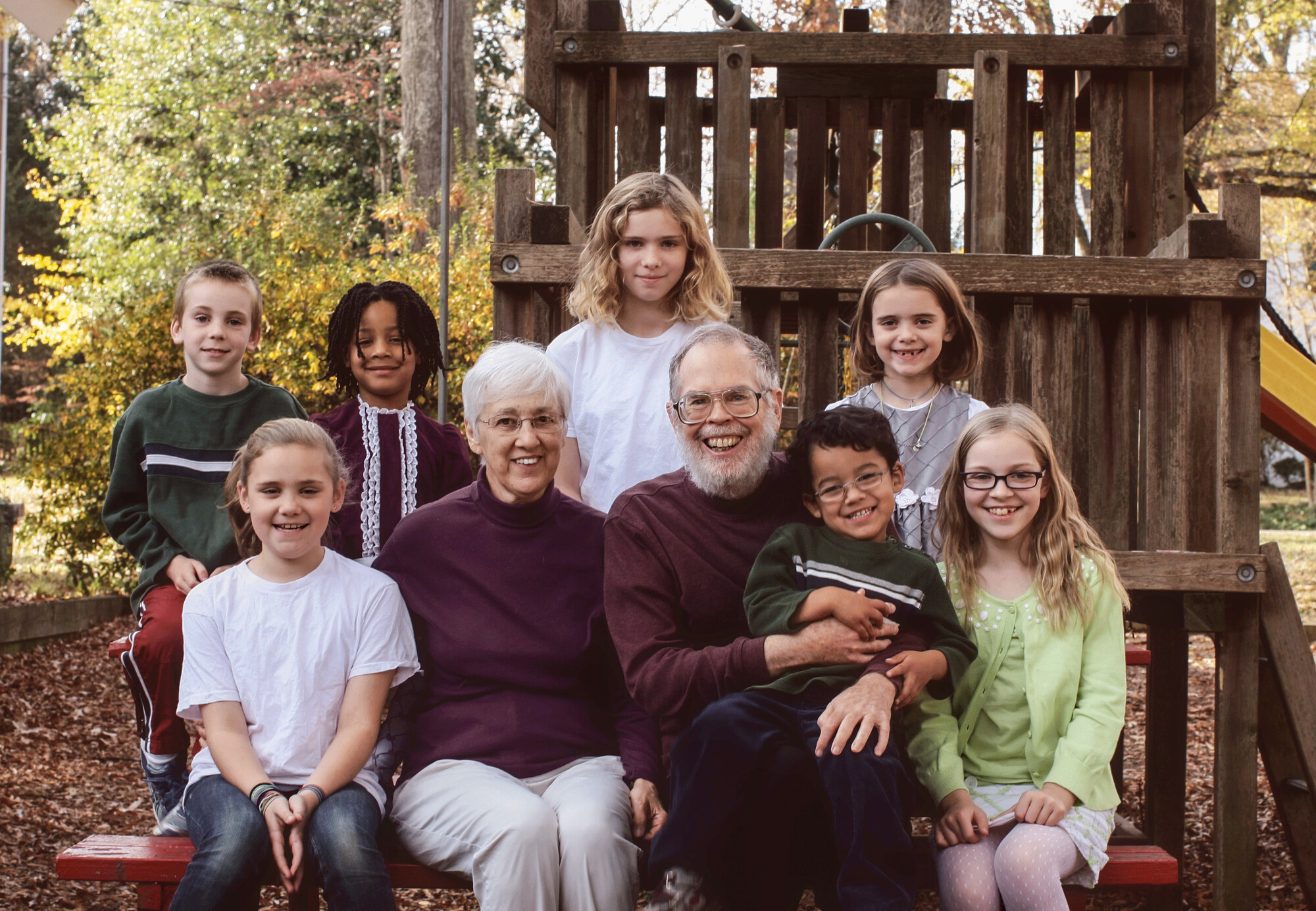 A group of two adults and seven children pose together, smiling on a wooden play structure in an outdoor setting with trees in the background.