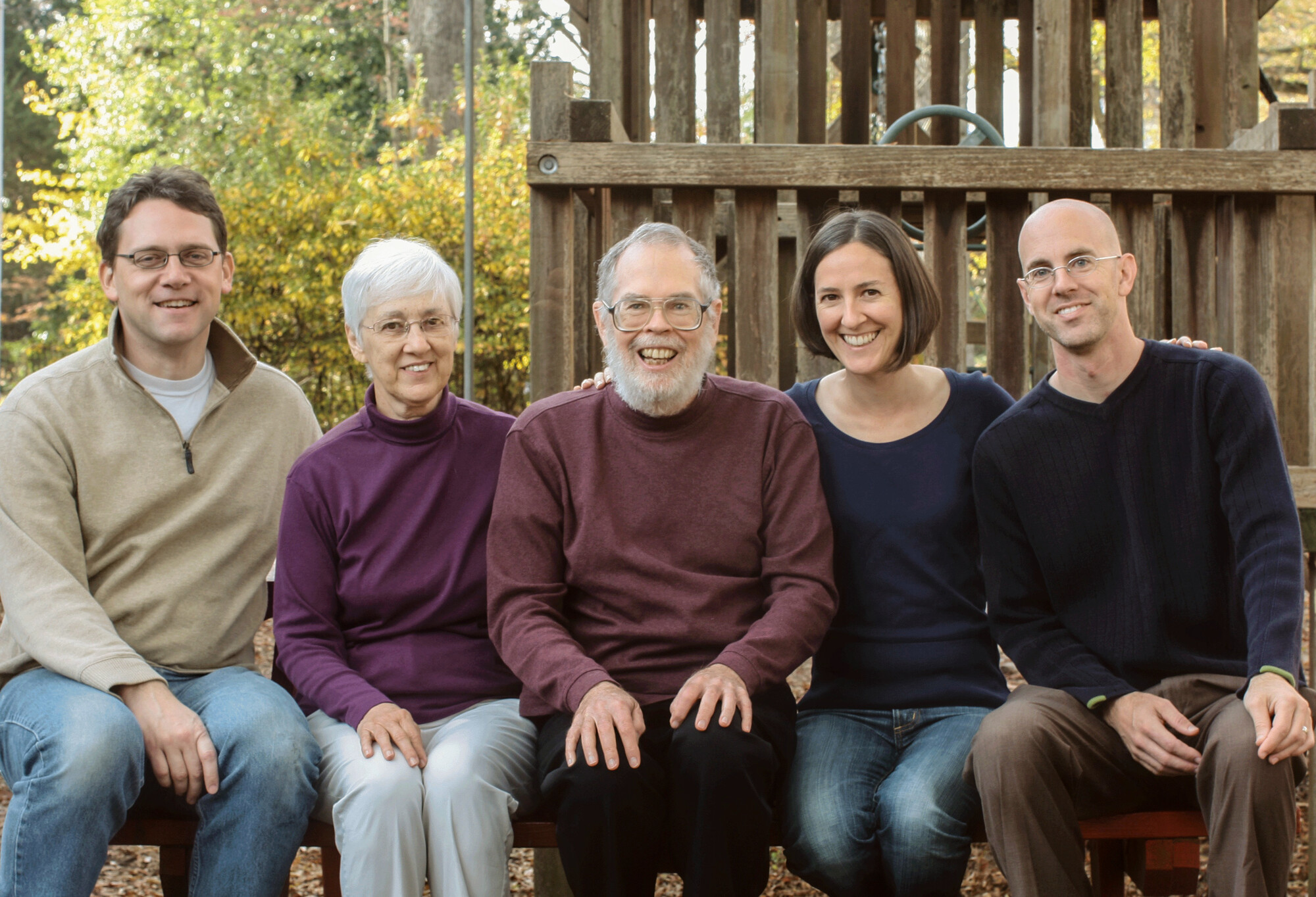 A family of five adults is seated outdoors, smiling at the camera. They are in front of a wooden play structure, surrounded by trees. The setting suggests a calm, autumn day.