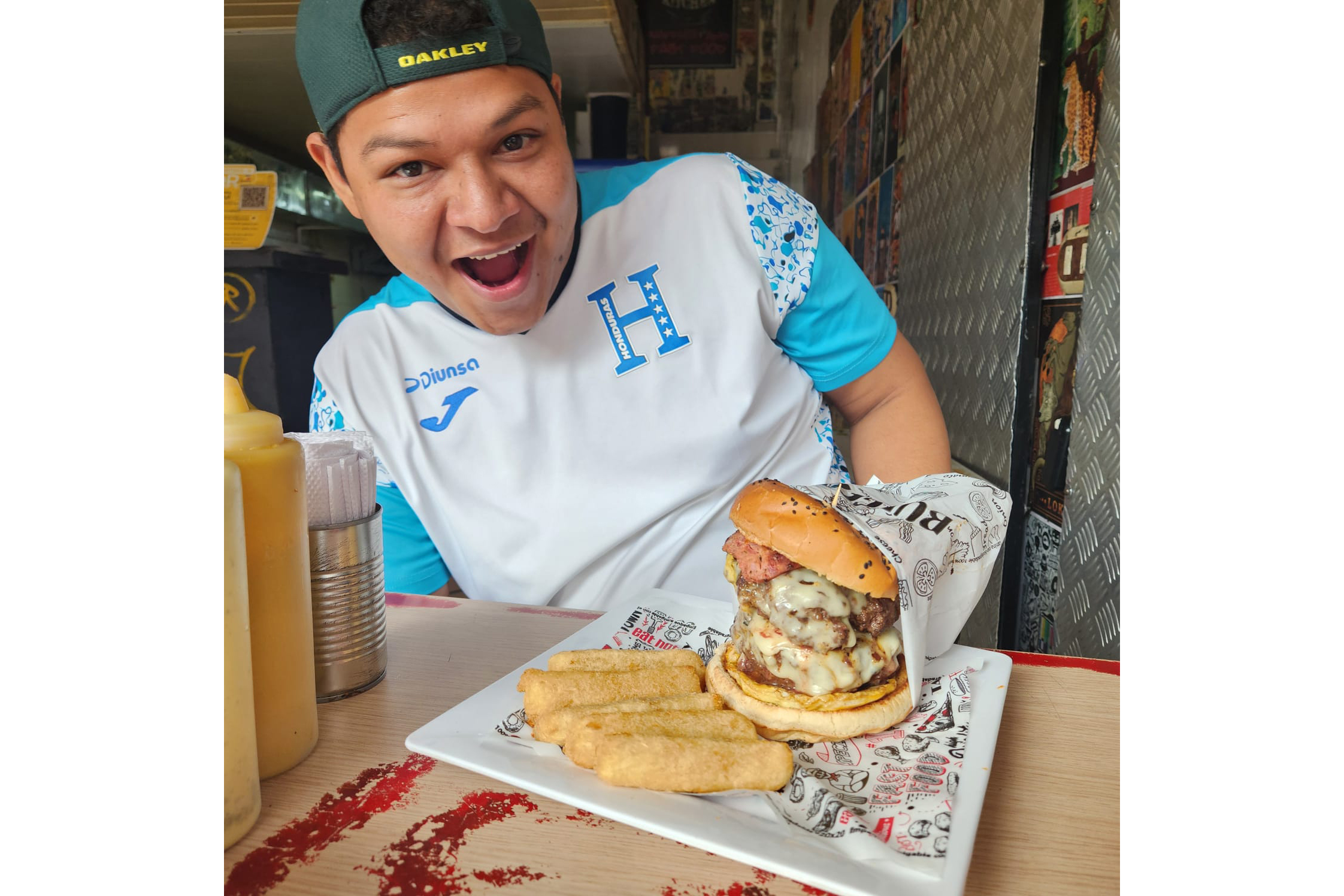 A young man poses with a large cheeseburger at a table