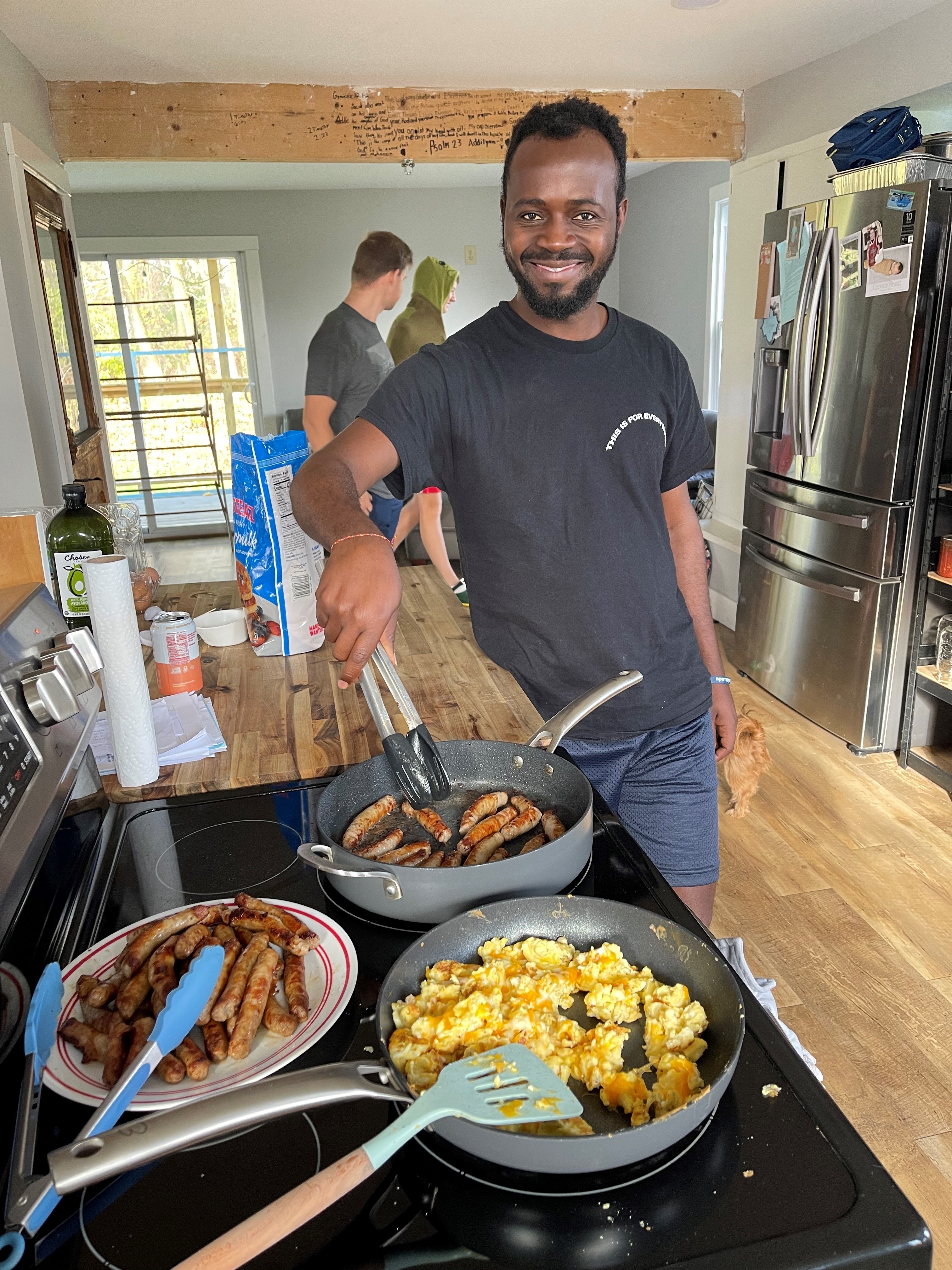 IVEP participant stands smiling and holding tongs in front of a stove cooking sausages and eggs.